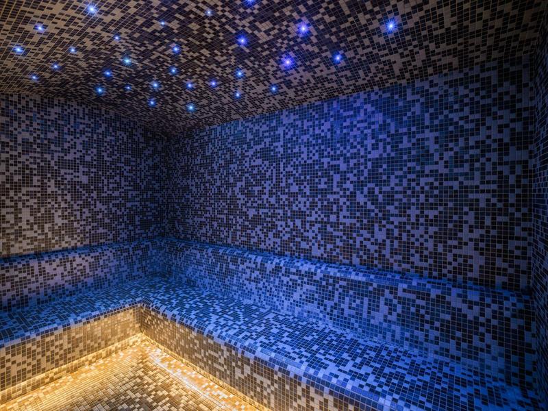 Sauna moderno con pareti e panchine in mosaico blu e illuminazione soffusa.