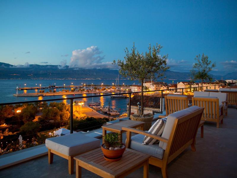 Avondterras met houten meubels met uitzicht op verlichte haven en bergen.