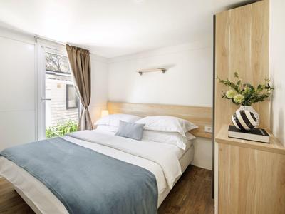 Chambre lumineuse avec lit double, linge blanc et couverture bleue près de la fenêtre avec rideaux.