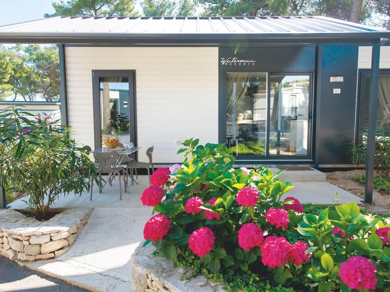 Maison de vacances moderne avec terrasse, entourée de fleurs roses éclatantes et d'arbres.