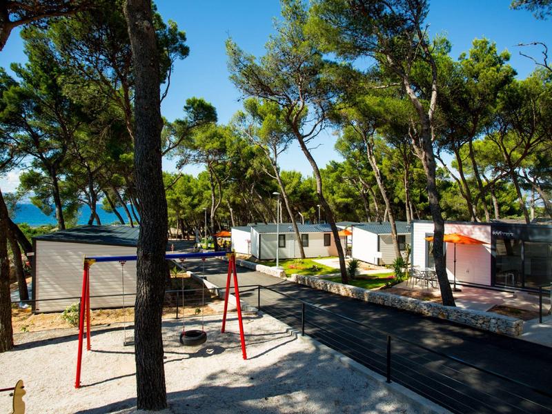 Aire de jeux avec balançoire entre des pins dans un parc de vacances au bord de la mer.