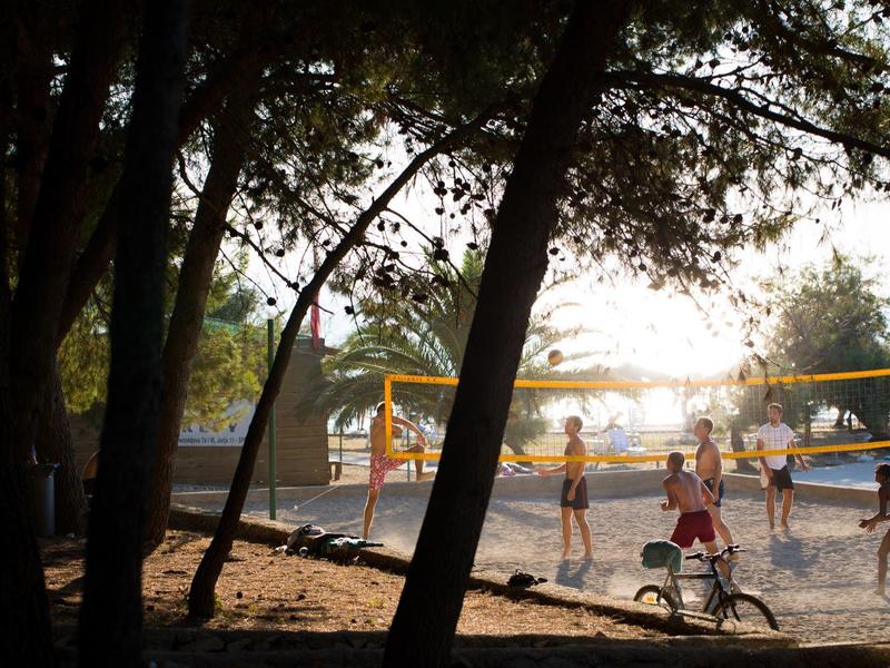 Bambini che giocano a pallavolo su un campo sabbioso al sole tra gli alberi.
