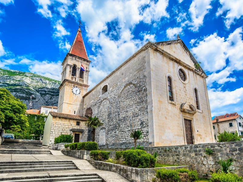 Historische Steinkirche mit rotspitzigem Turm vor blauem Himmel und grünen Bergen.