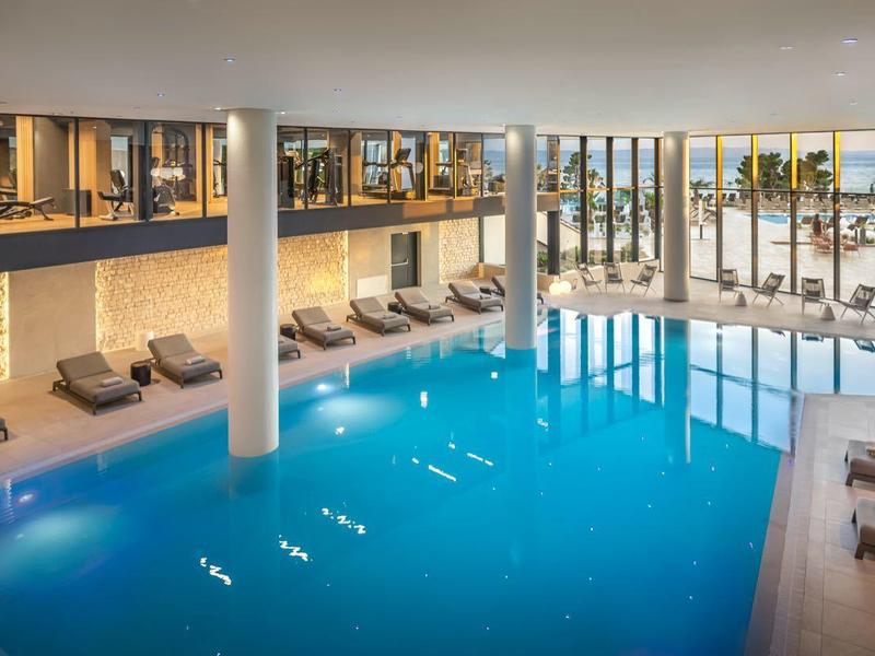 Moderner Innenpool mit großen Fenstern, blauer Wasserfläche, Liegen und warmem Lichtambiente.