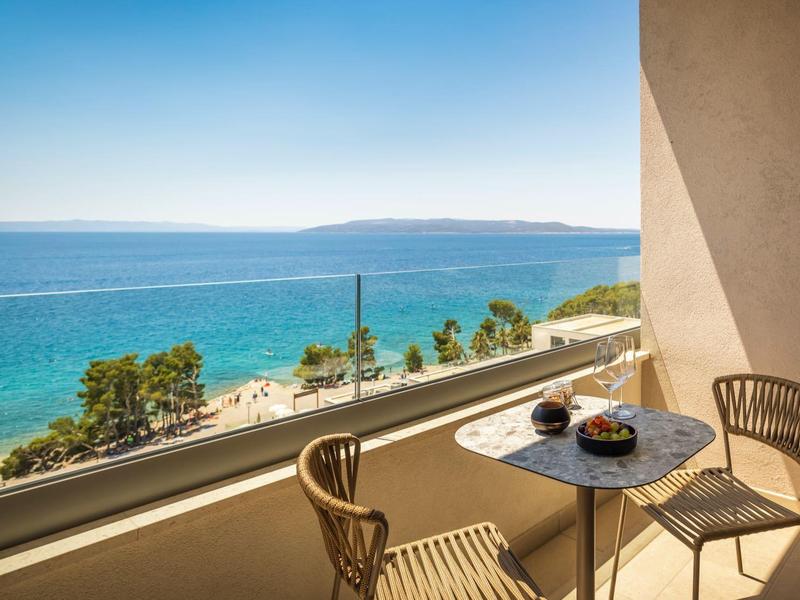 Balkon mit kleinem runden Tisch, zwei Stühlen, Obstschale und Meerblick bei klarem Himmel.