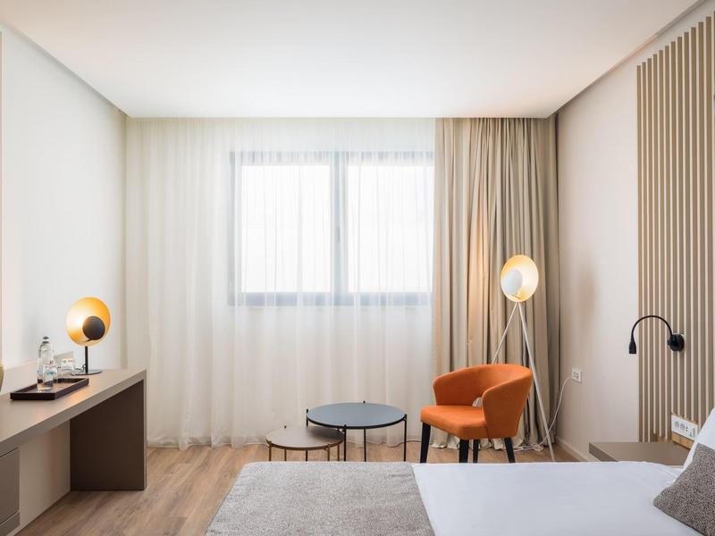 Hell eingerichtetes Schlafzimmer mit orangefarbenem Sessel, runden Tischen und großem Fenster.