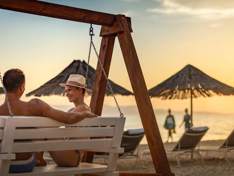 Zwei Personen sitzen auf einer Holzschaukel am Strand bei Sonnenuntergang, Palmen und Sonnenschirme im Hintergrund.