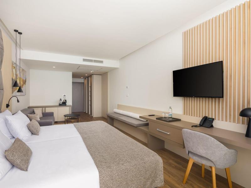 Modernes Hotelzimmer mit großem Bett, braunen und weißen Farbtönen, Wand-TV und Schreibtischbereich.