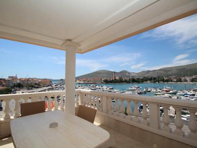 Balcony with white table and chairs overlooking a harbor with boats and mountains in the background.