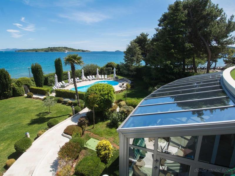 View of a hotel area with pool, lawn, palm trees, and sea in the background.