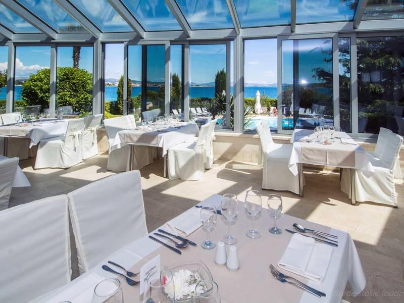 Bright restaurant with glass roof and white set tables, overlooking pool and garden.