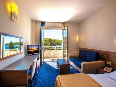 Hotelkamer met balkon, zithoek, tv en uitzicht op een beboste heuvel.