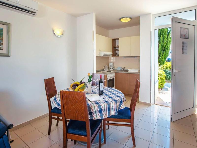 Jadalnia z niebiesko-białym obrusem i krzesłami, otwarte drzwi na zewnątrz, jasny kącik kuchenny.