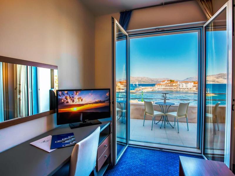 Pokój hotelowy z biurkiem, krzesłem, telewizorem i otwartymi drzwiami na balkon z widokiem na morze.