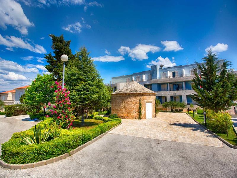 Mały kamienny budynek i ogród z drzewami przed nowoczesnym hotelem pod niebem z błękitnymi chmurami