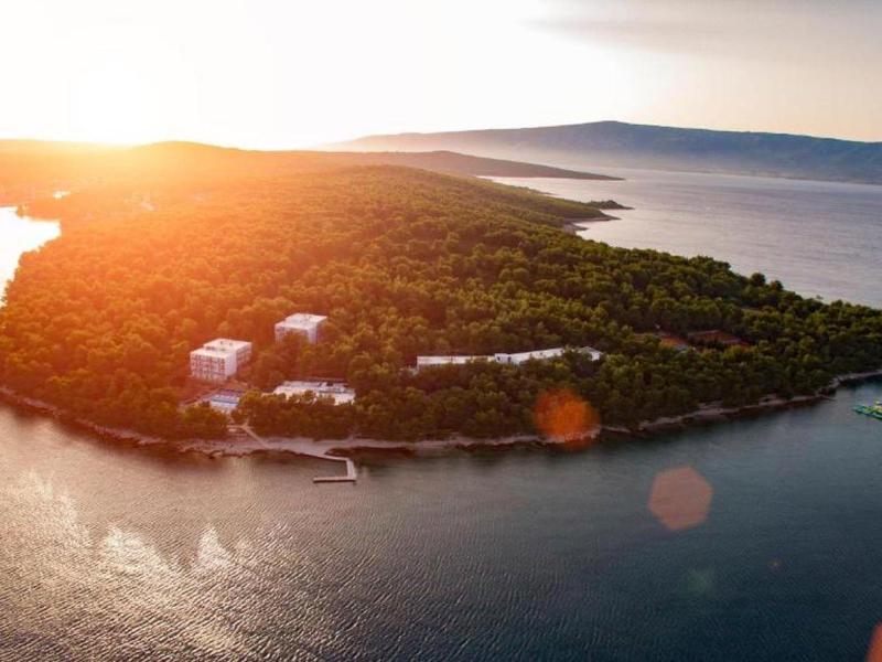 Západ slunce nad zalesněným ostrovem s hotely a loděmi na klidné vodě.