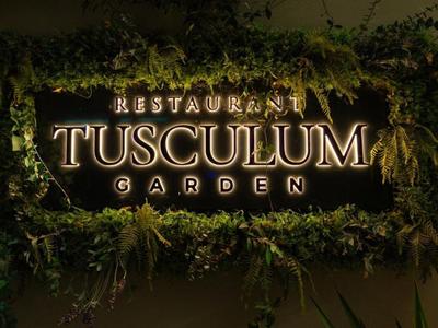 Beleuchteter Restaurant-Schild 'TUSCULUM GARDEN' mit grünem Pflanzenrahmen.