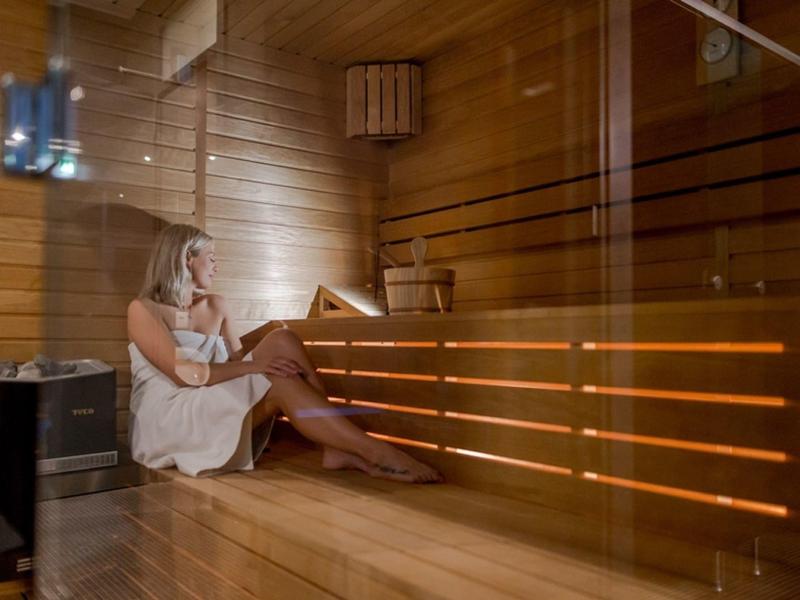 Frau entspannt in Sauna mit Holzwänden und Bänken, sitzt in weißem Handtuch.