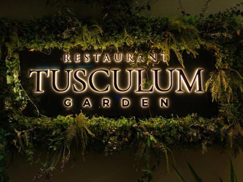 Beleuchteter Restaurant-Schild 'TUSCULUM GARDEN' mit grünem Pflanzenrahmen.