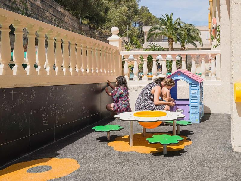 Kinderen spelen op een kleurrijk speeltoestel in een binnenplaats met hoge muren en palmbomen op de achtergrond.