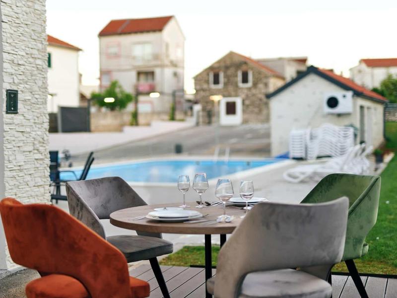 Gezellige stoelen rond een gedekte tafel op een terras met zwembad en huizen op de achtergrond.