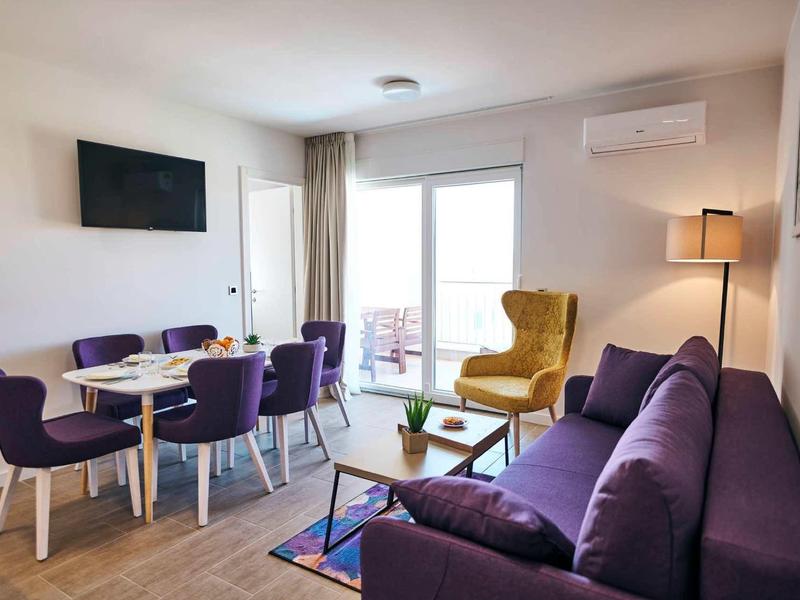 Modern ingerichte woonkamer met paarse bank, gele fauteuil en kleine eethoek bij balkon.