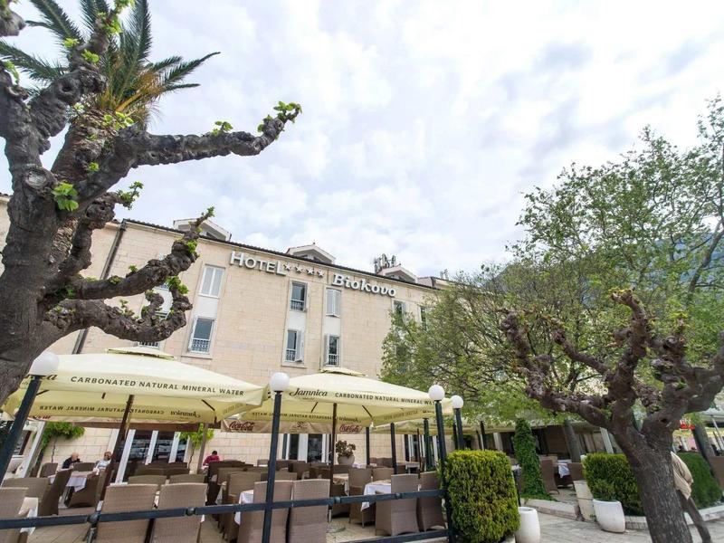 Gebäude mit Café-Terrasse, Sonnenschirmen, blühenden und beschnittenen Bäumen, bewölkter Himmel.