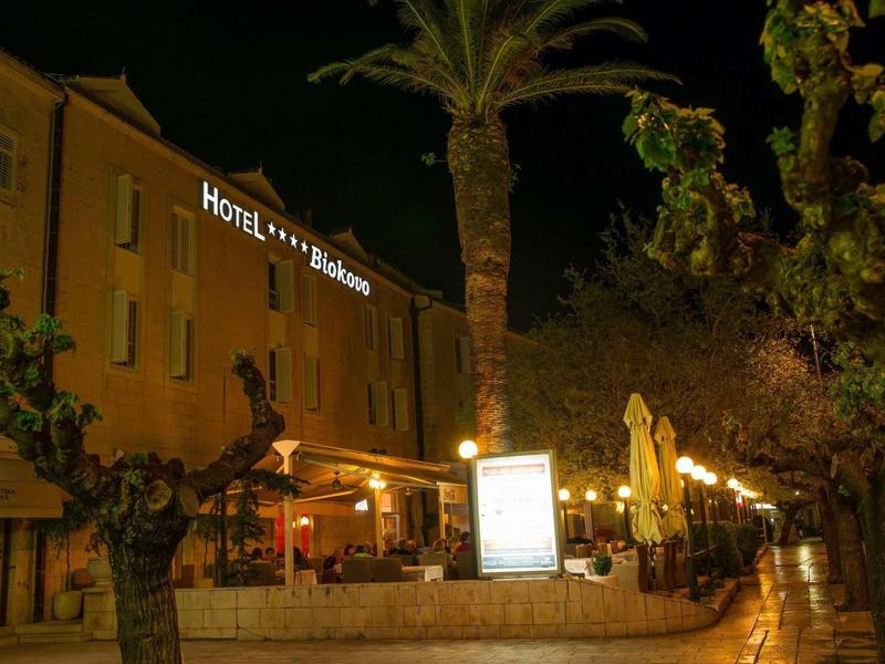 Hotelgebäude mit beleuchtetem Schriftzug, Terrassenbeleuchtung, Palmen und Bäumen bei Nacht.