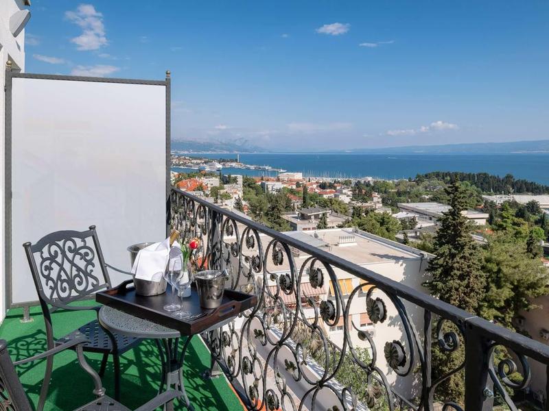 Balkon z ozdobną balustradą, dwoma zielonymi krzesłami i widokiem na miasto i morze.