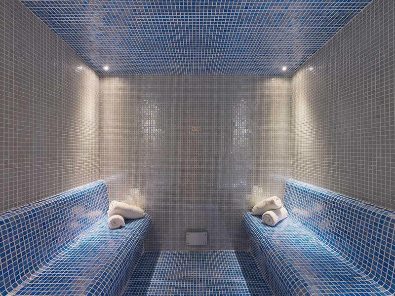 Luksusowa łaźnia parowa z niebieskimi kafelkami i zwiniętymi ręcznikami na ławkach.