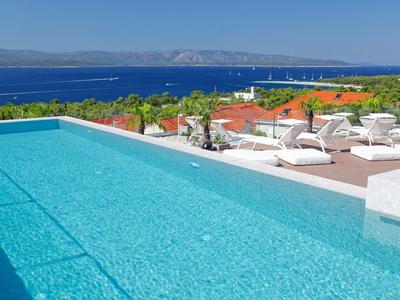Infinity-Pool mit klarem Wasser, Liegestühlen und Blick auf das blaue Meer und rote Dächer.