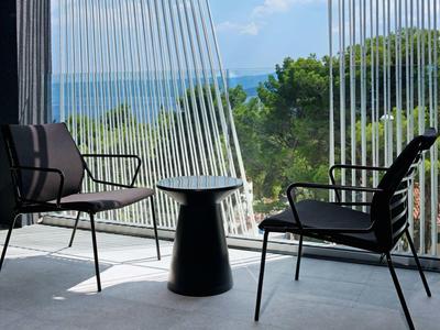 Balkon mit zwei Stühlen und kleinem Tisch mit Blick auf Bäume und Meer bei klarem Himmel.