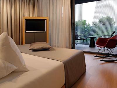 Modern eingerichtetes Hotelzimmer mit Bett, Fernseher und Balkon mit Stühlen und Tisch.