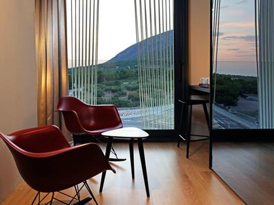 Warmer Innenraum mit zwei roten Sesseln, kleinem Tisch und großer Glasfront mit Blick auf Hügel bei Sonnenuntergang.