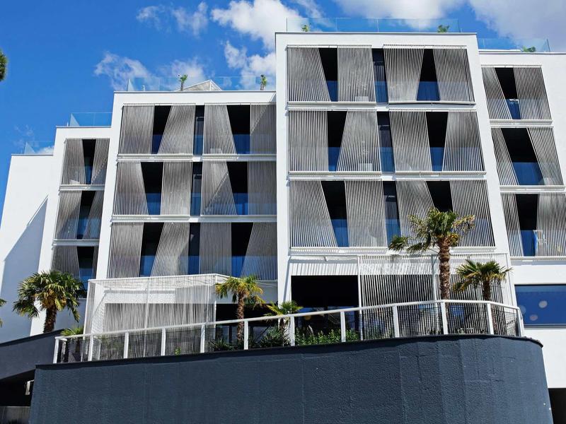 Modernes, weißes Gebäude mit Glasbalkonen unter blauem Himmel und kleinen Palmen im Vorgarten.