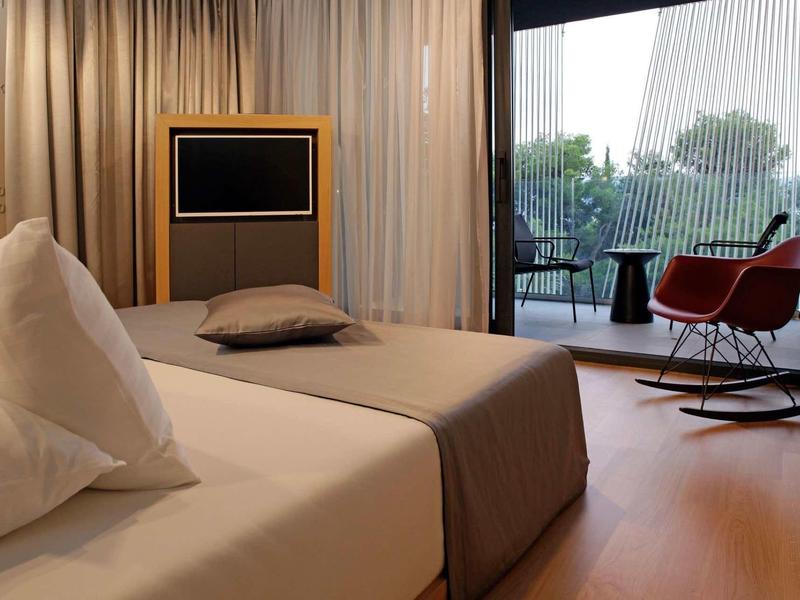 Modern eingerichtetes Hotelzimmer mit Bett, Fernseher und Balkon mit Stühlen und Tisch.