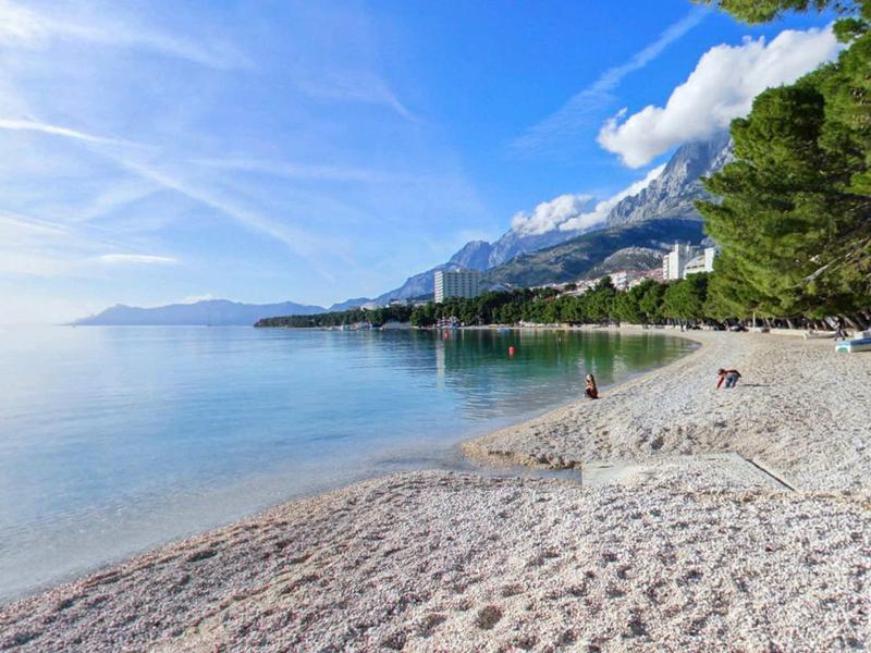 Beach with clear water, mountains in the background, and hotel buildings under a blue sky.