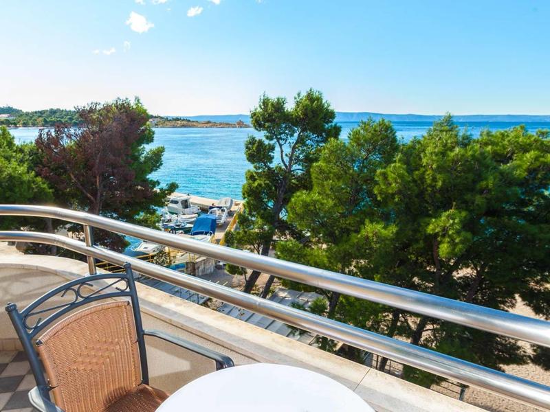 View from a balcony with metal railing overlooking the sea and trees under a clear sky.