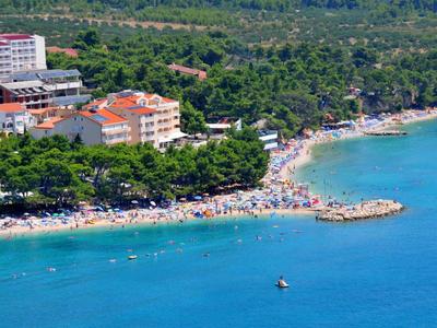 Spiaggia affollata con ombrelloni accanto a hotel lungo una costa boscosa.