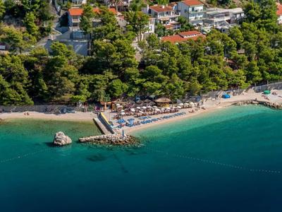 Spiaggia con lettini, ombrelloni e acqua limpida, circondata da alberi verdi e edifici.