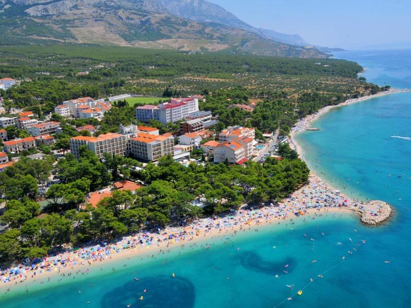 Veduta aerea di una spiaggia affollata con acqua turchese e hotel sullo sfondo verde.