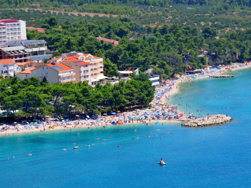 Spiaggia affollata con ombrelloni accanto a hotel lungo una costa boscosa.