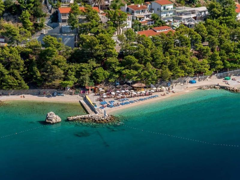 Spiaggia con lettini, ombrelloni e acqua limpida, circondata da alberi verdi e edifici.