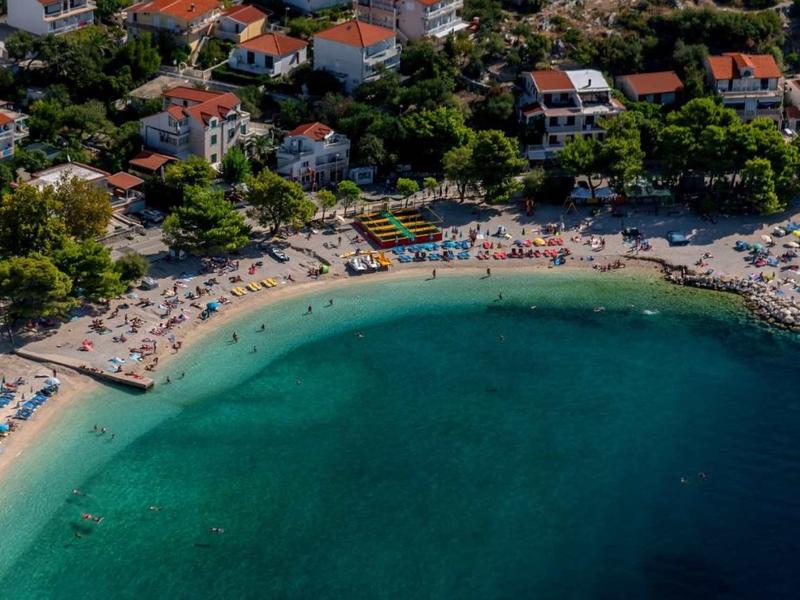 Piccola spiaggia con acqua limpida, circondata da case e vegetazione rigogliosa.