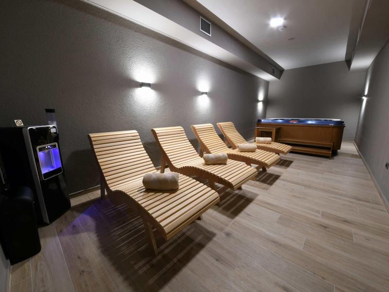 Area relax moderna con quattro lettini in legno, asciugamani e vasca idromassaggio con luce soffusa.