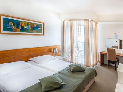 Habitación de hotel luminosa con cama doble, escritorio, silla y grandes ventanas con cortinas.