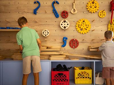 Two boys play at a wooden wall with various mechanical parts in a play area.