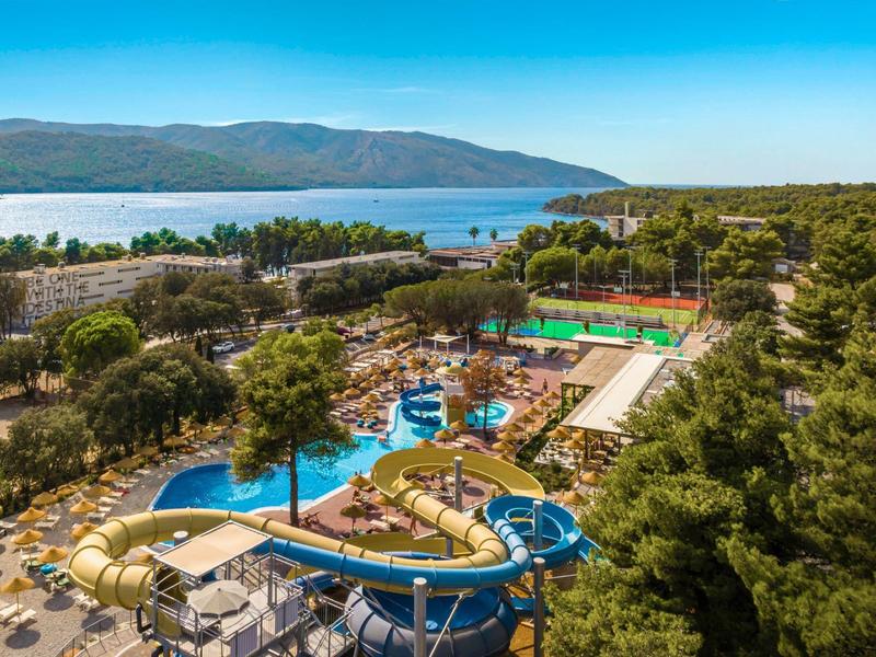 View of hotel pool with water slides, sun umbrellas, and pine trees with sea in background.