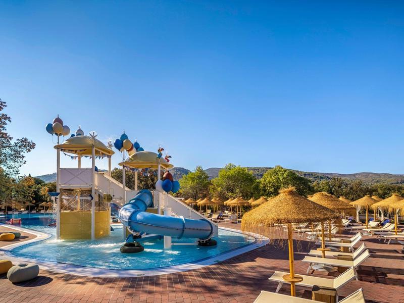Shallow pool with water slides and lounge chairs under sunshades on a sunny day.