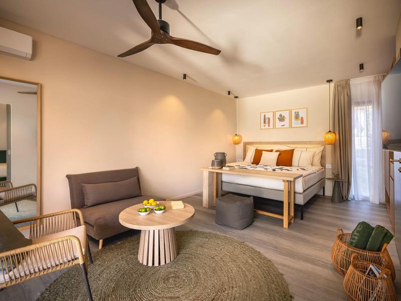 Bright hotel room with double bed, seating area, ceiling fan, and wooden floor.